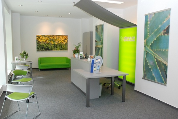 Modernes Hörakustiker-Fachgeschäft mit Empfangstresen, grüner Couch und Pflanzen.